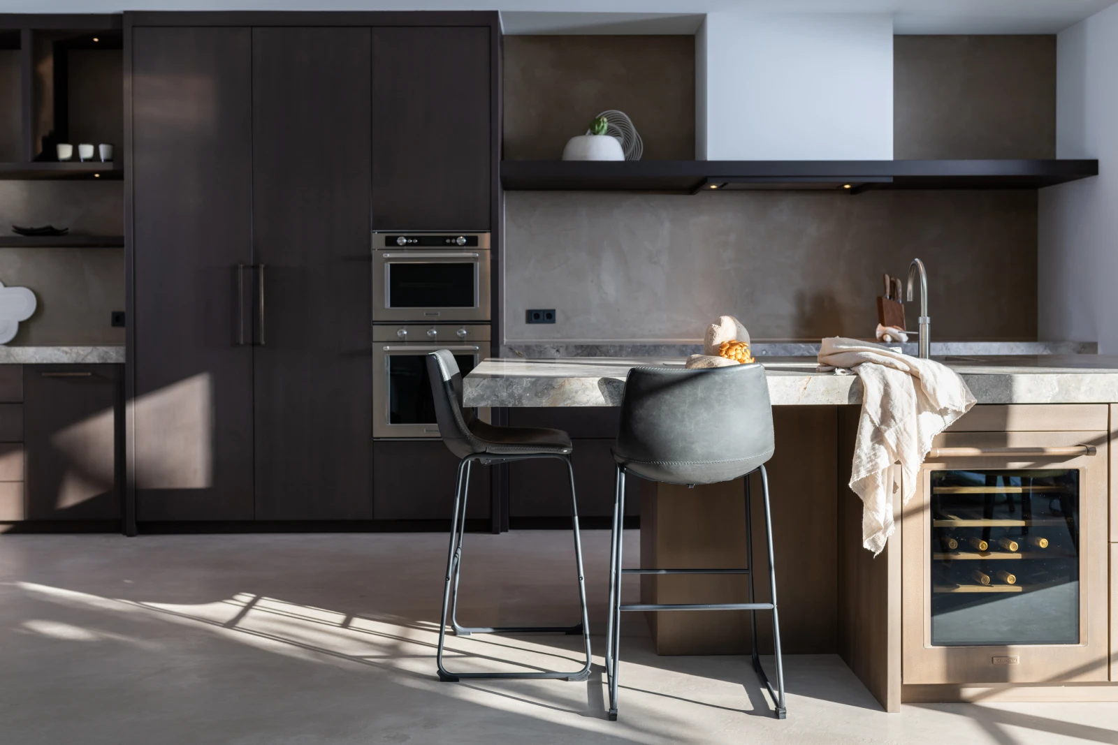 Moderne keuken met donkere kasten, ingebouwde ovens, een marmeren keukenblad eiland met barkrukken, een gootsteen en een wijnkoeler. Een beige handdoek en een brood liggen op het aanrecht, met planken en minimale decoratie op de achtergrond.