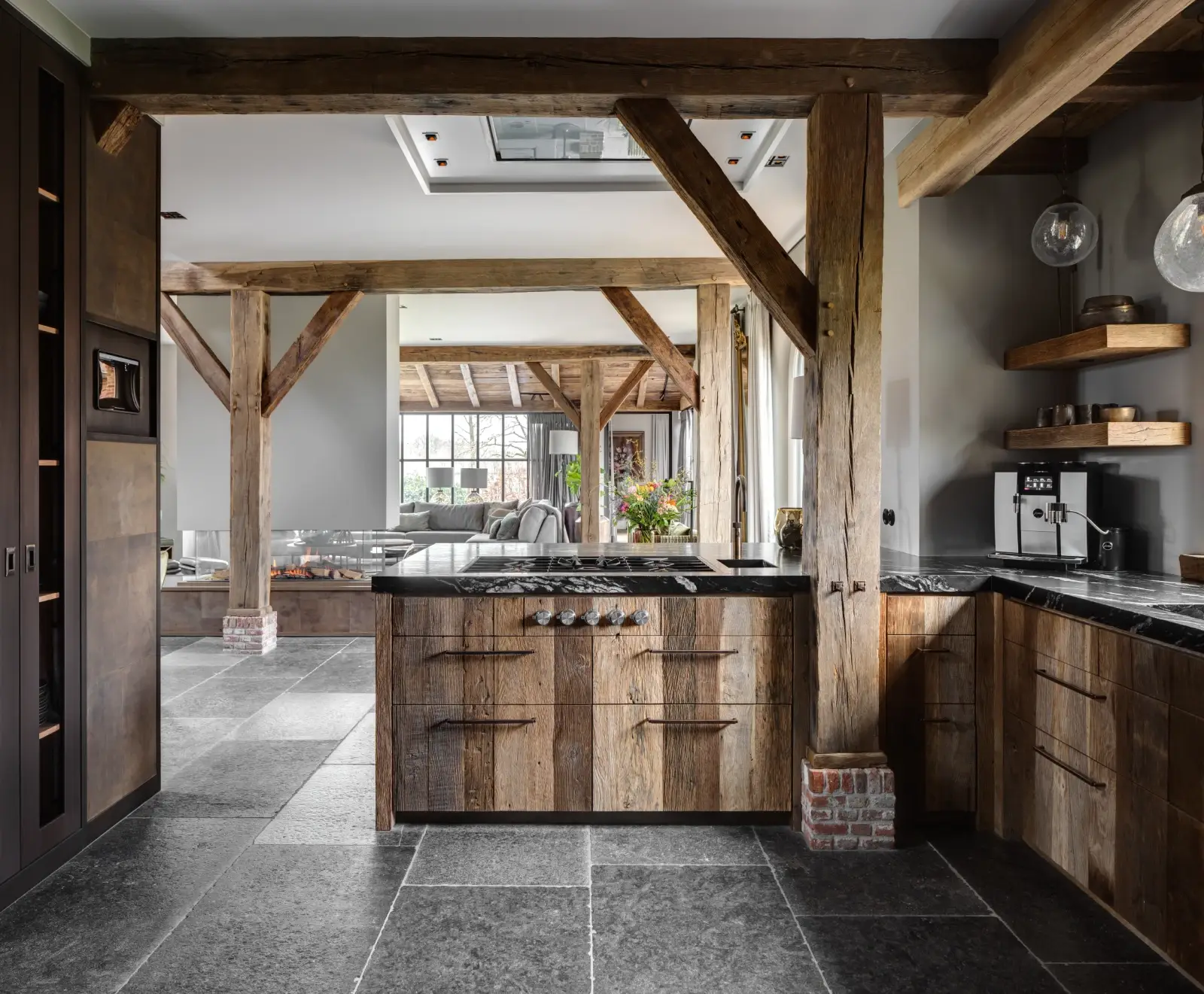 Rustieke keuken met houten balken, stenen tegelvloer, donkerhouten kasten en een oud eiken keukengevoel. Voorzien van een granieten keukenblad, open planken en grote ramen voor natuurlijk licht. Gezellige woonkamer met banken op de achtergrond.